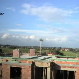 Wznoszenie ścian z czerwonej cegły na budowie domu jednorodzinnego, widok na okolicę z polami i zabudowaniami, majówka na dachu.